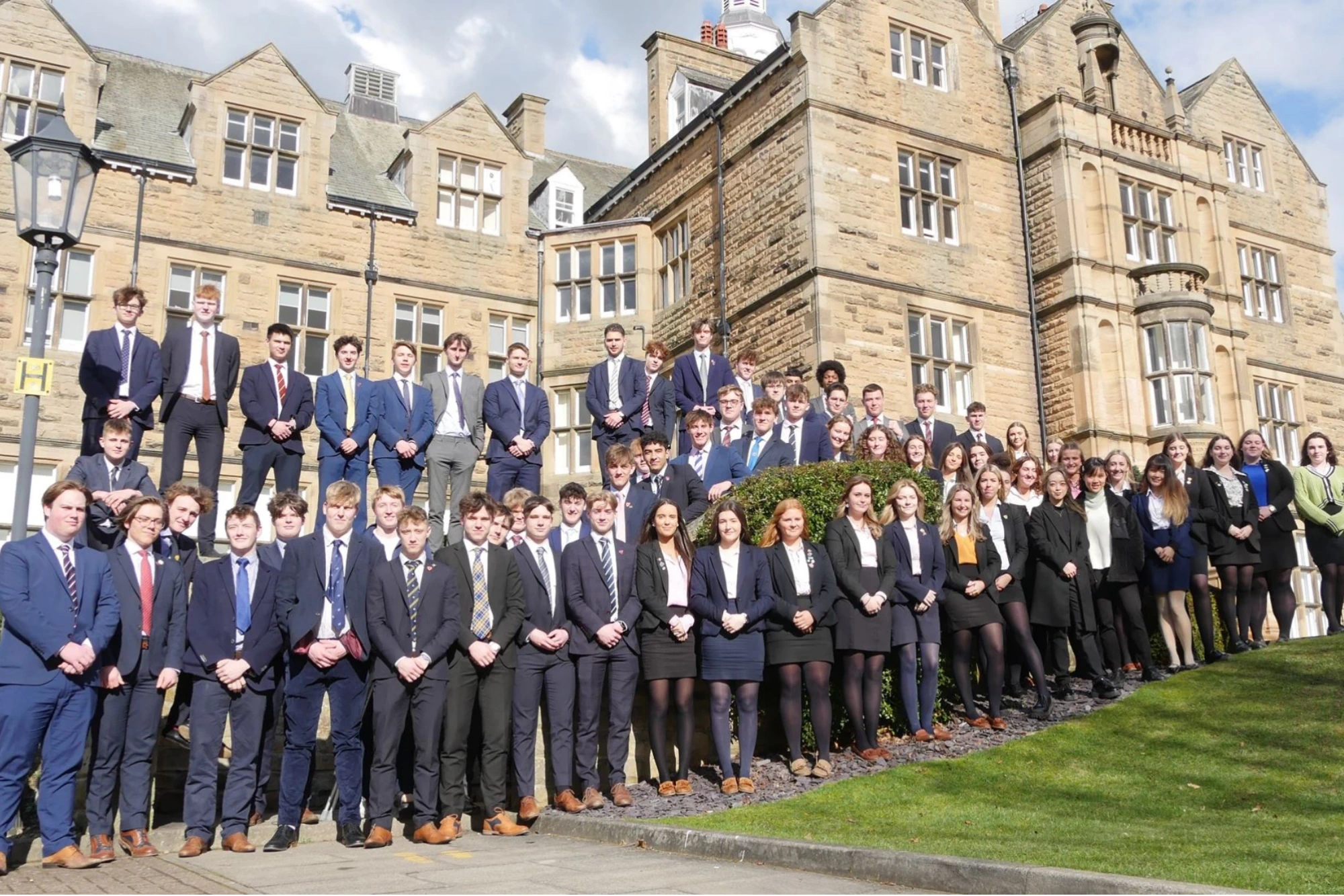 A large group of Barney students in formal wear pose outside a traditional stone school building on results day. They are arranged on steps and a grassy slope, with a partly cloudy sky overhead.