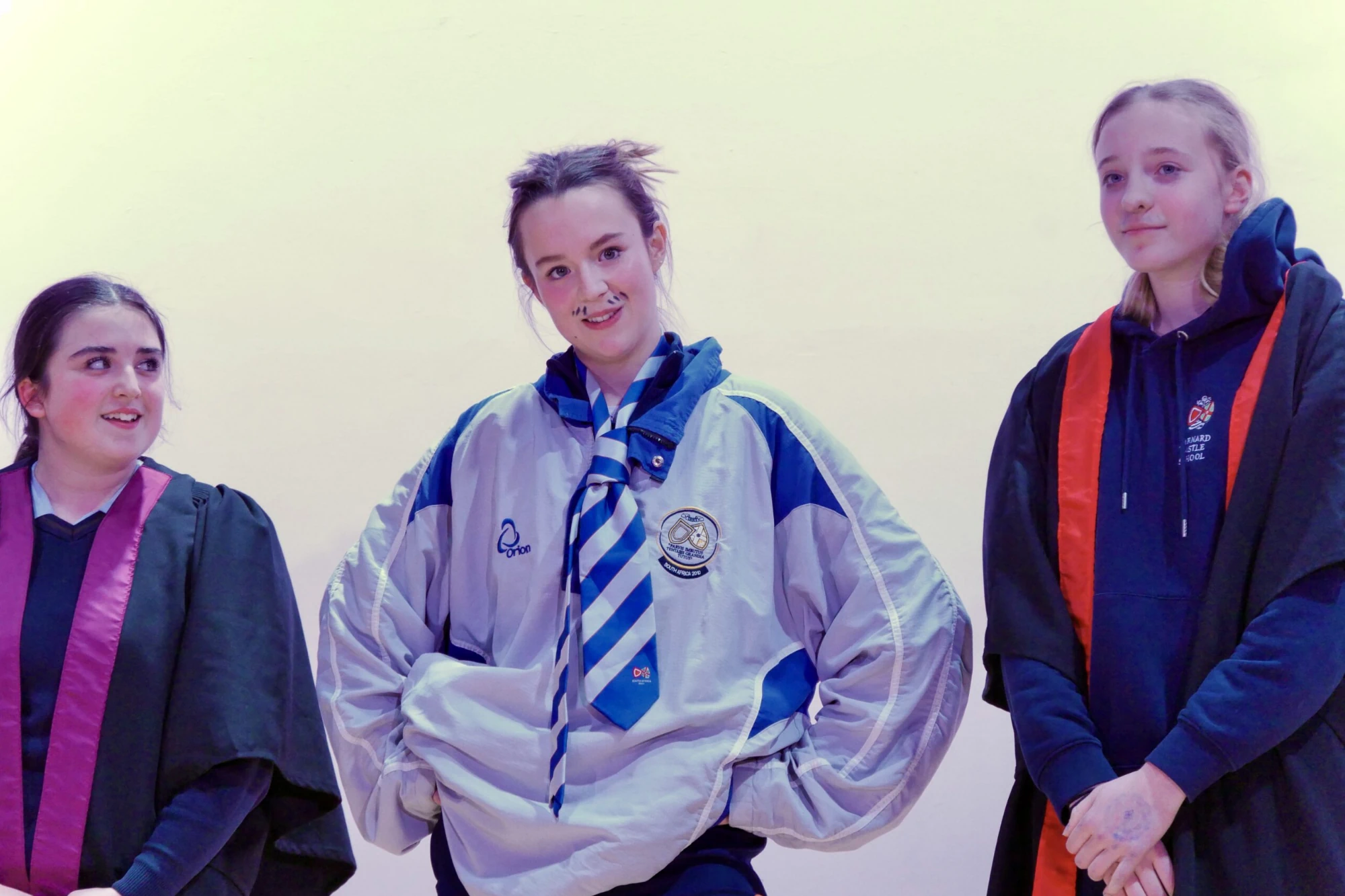 Three students from Barnard Castle School on stage for House Drama 2024. The middle student wears an athletic jacket and tie with theatrical makeup, including a drawn-on mustache.