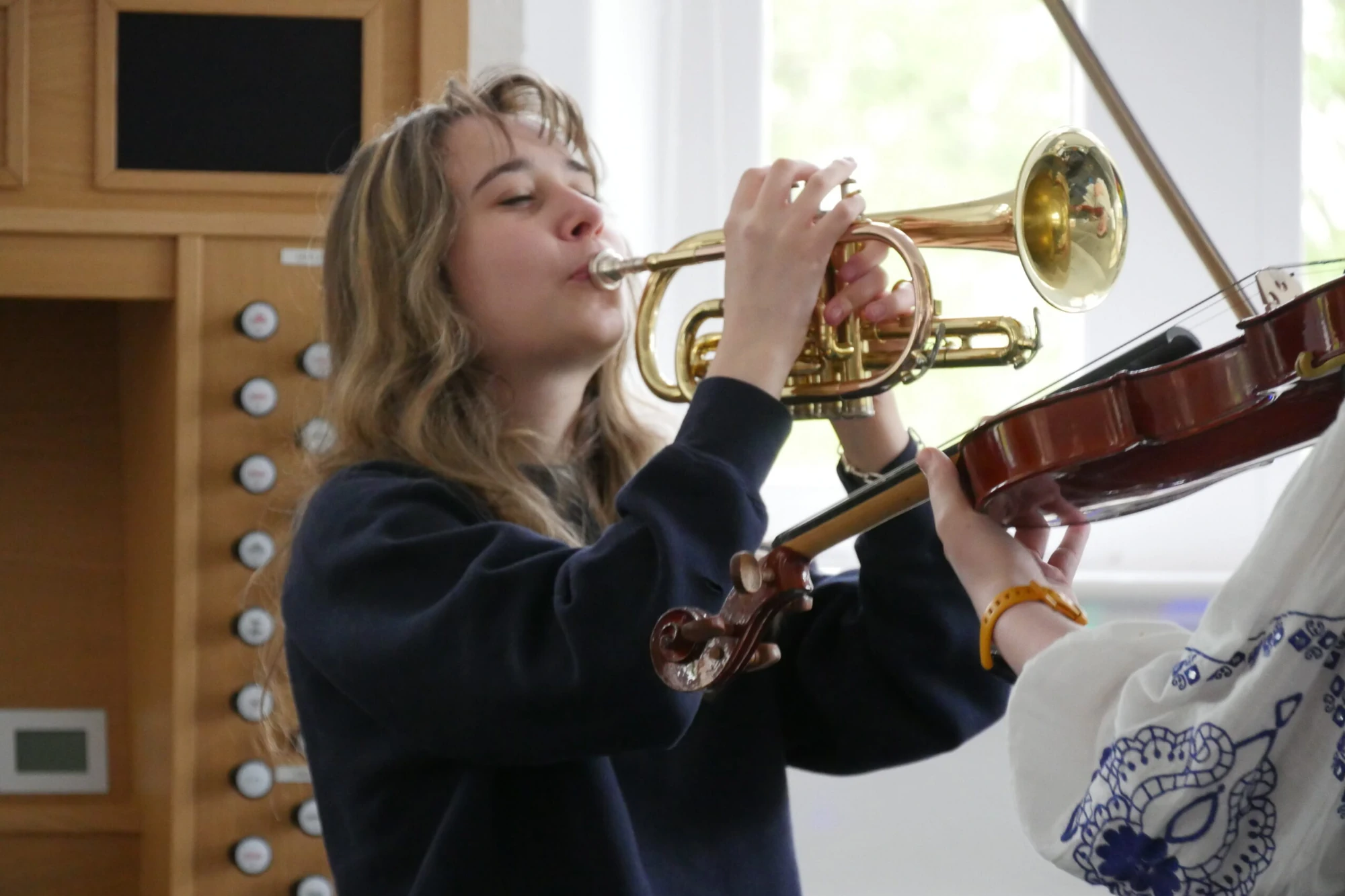 A close-up shot captures a student with long, wavy brown hair playing a trumpet.