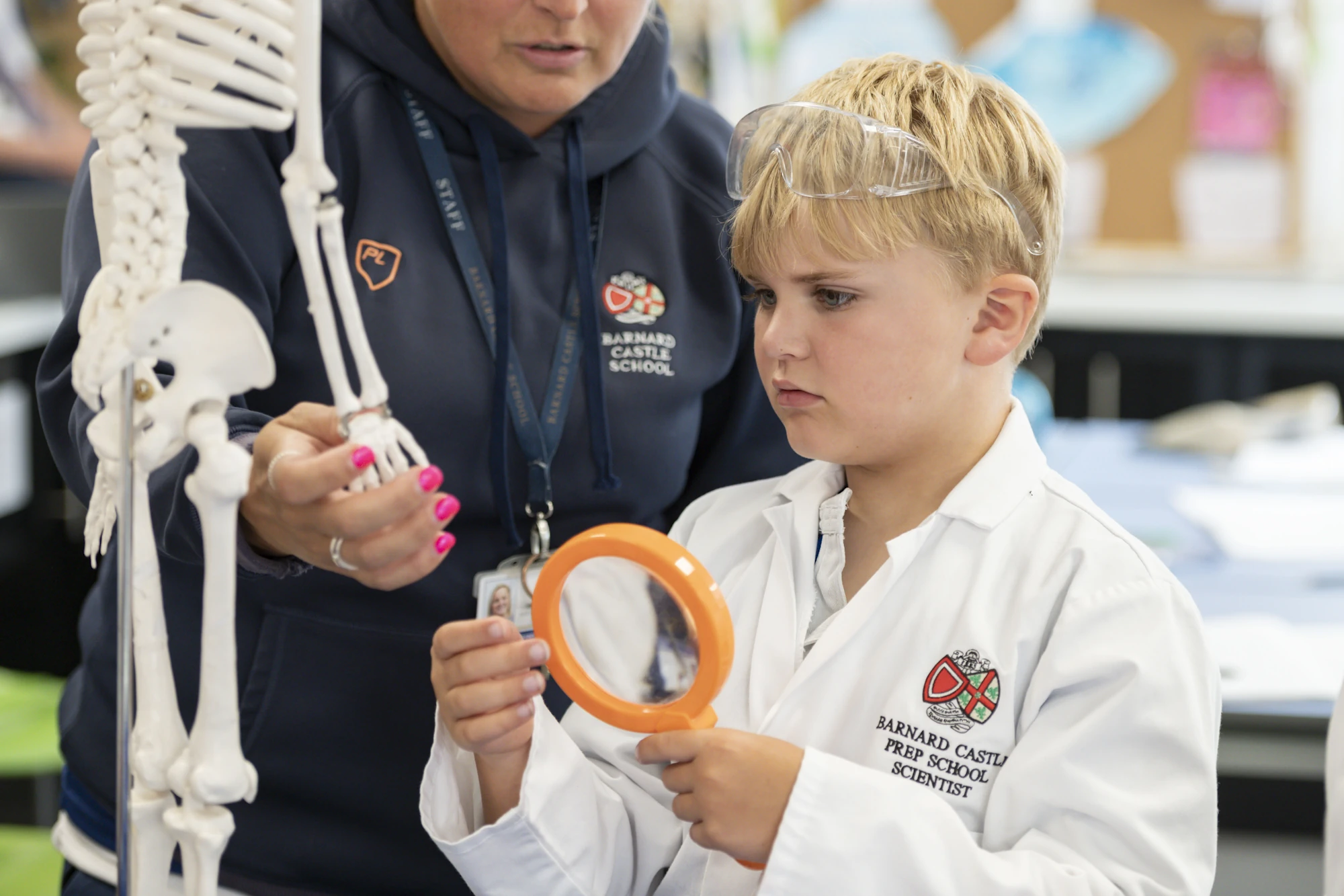 A young Barnard Castle School pupil in a lab coat, holding a magnifying glass with a teacher in the background.