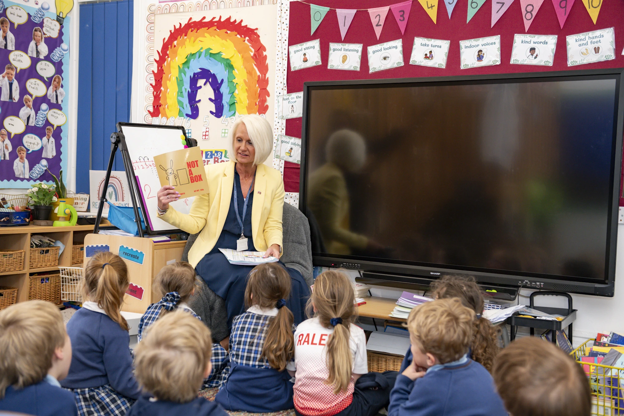 A Barnard Castle School teacher holding a book, in front of a class of Preppies who are sat on the mat.