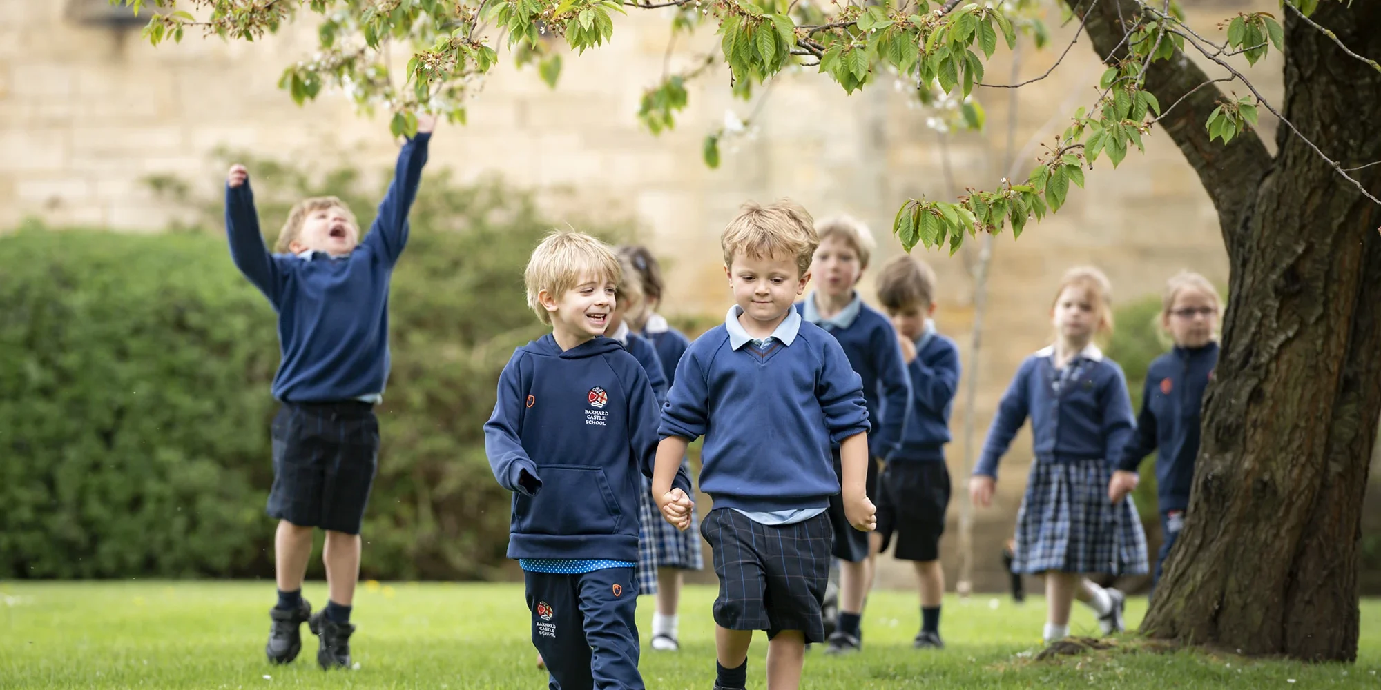 A group of Barnard Castle School pupils joyfully running together on a grassy field under a clear blue sky.