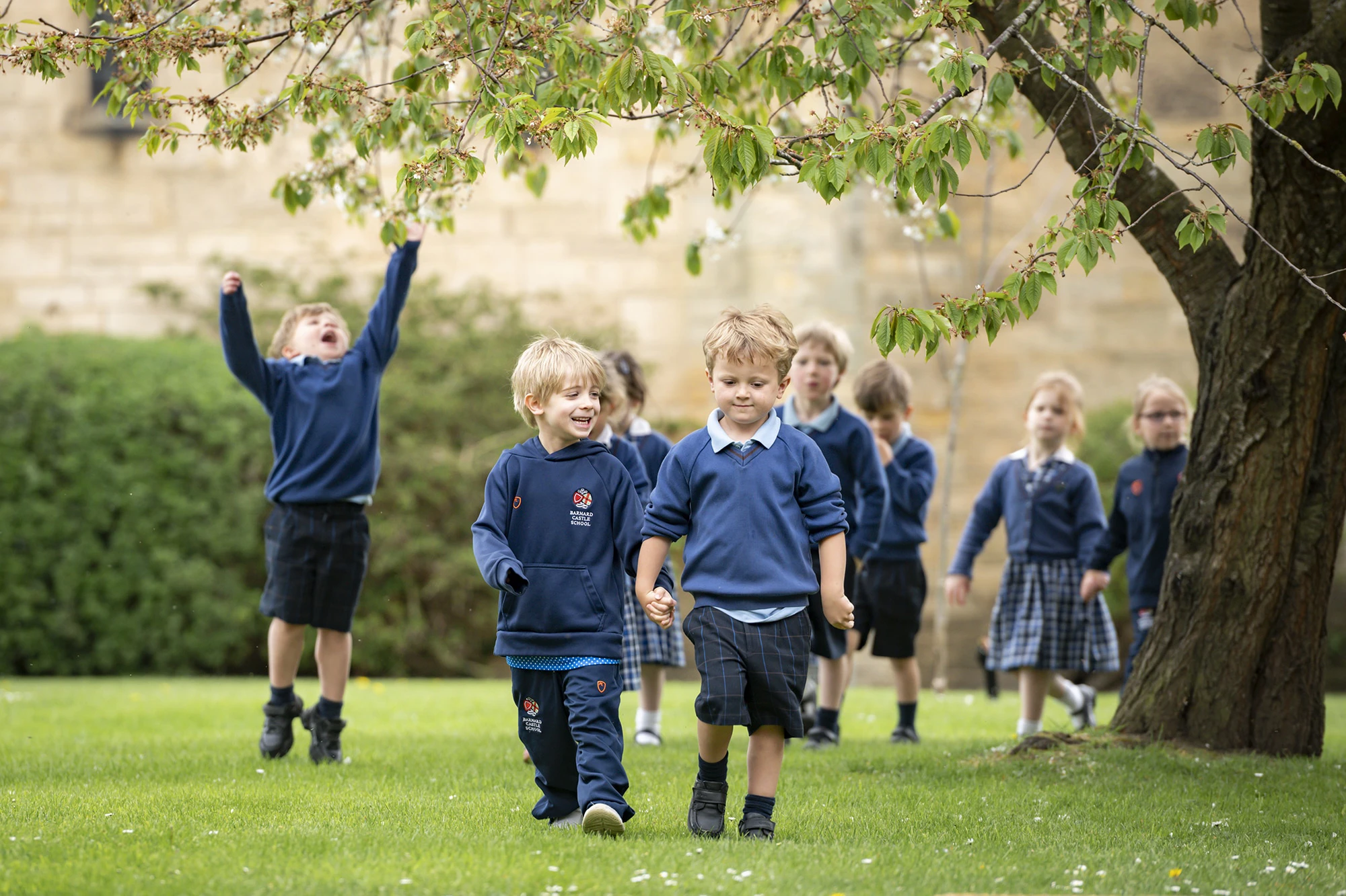 A group of Barnard Castle School pupils joyfully running together on a grassy field under a clear blue sky.
