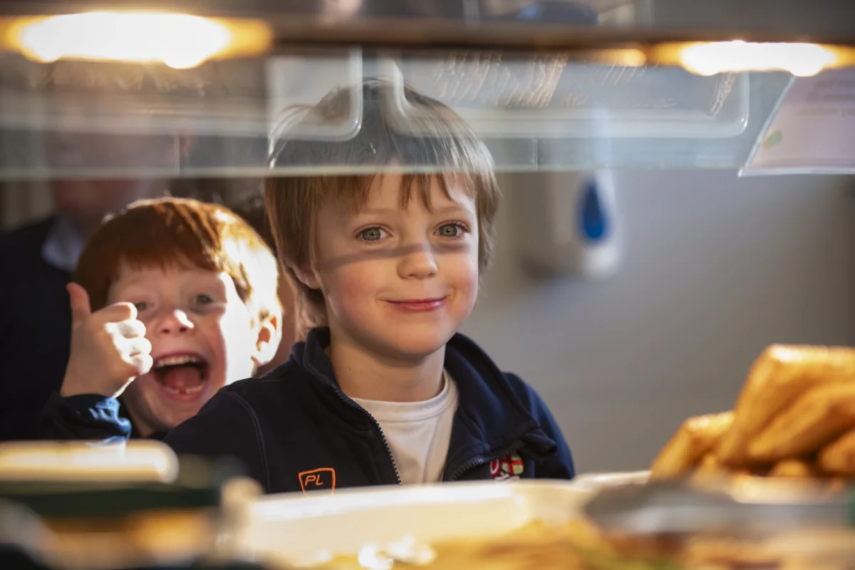 Two Barnard Castle School children in line in the dining hall, both smiling and one holding a thumbs up.