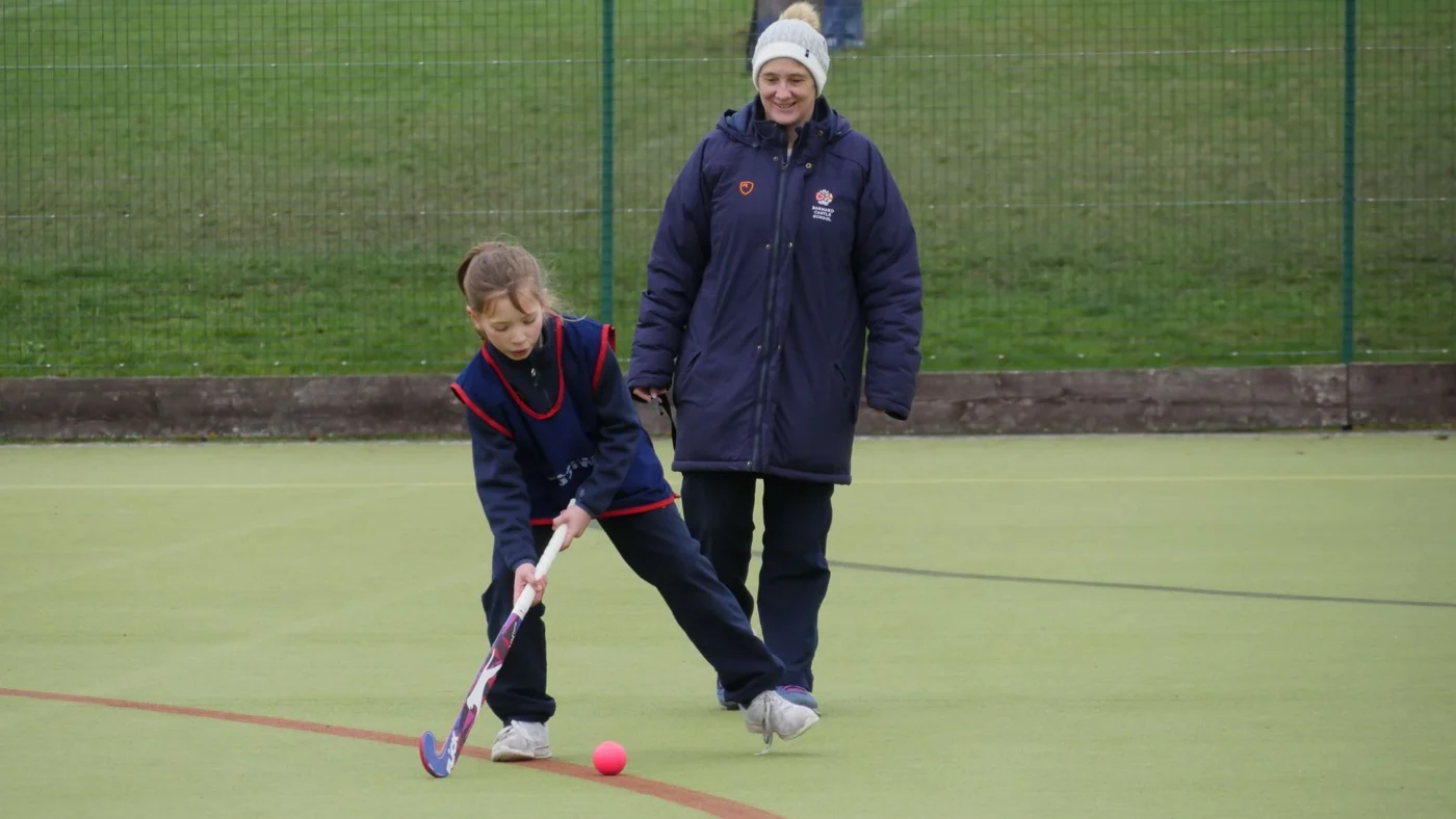 A Barnard Castle School student playing hockey with a teacher supervising.