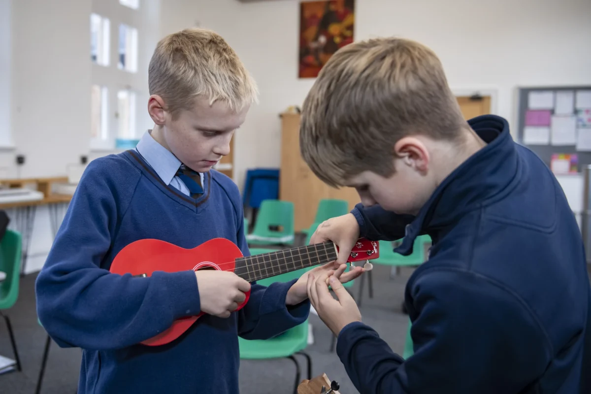 Two Barnard Castle School pupils playing ukuleles together in a bright classroom, one helping the other with chords.