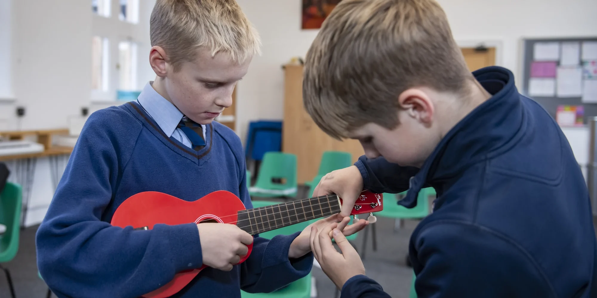 Two Barnard Castle School pupils playing ukuleles together in a bright classroom, one helping the other with chords.