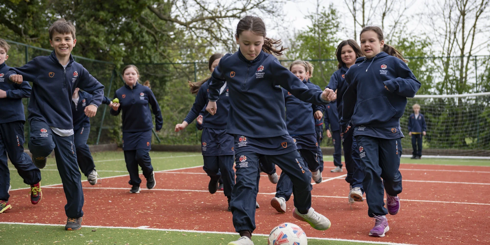 A group of young Barnard Castle School pupils joyfully playing football on a pitch, starring a girl kicking the ball.