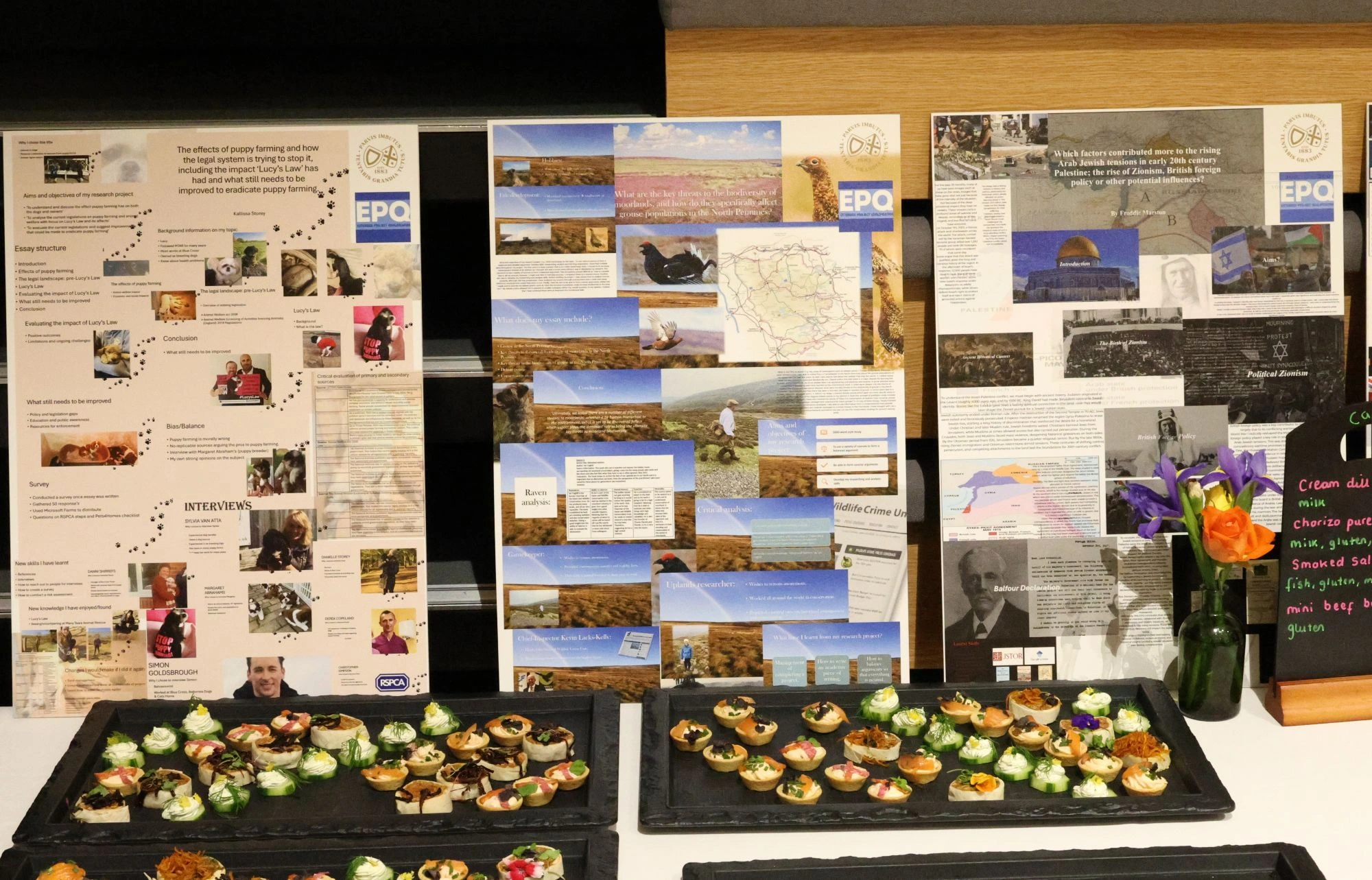 Three academic posters are displayed on a wall behind a table set with several trays of colourful appetisers and a small floral arrangement.