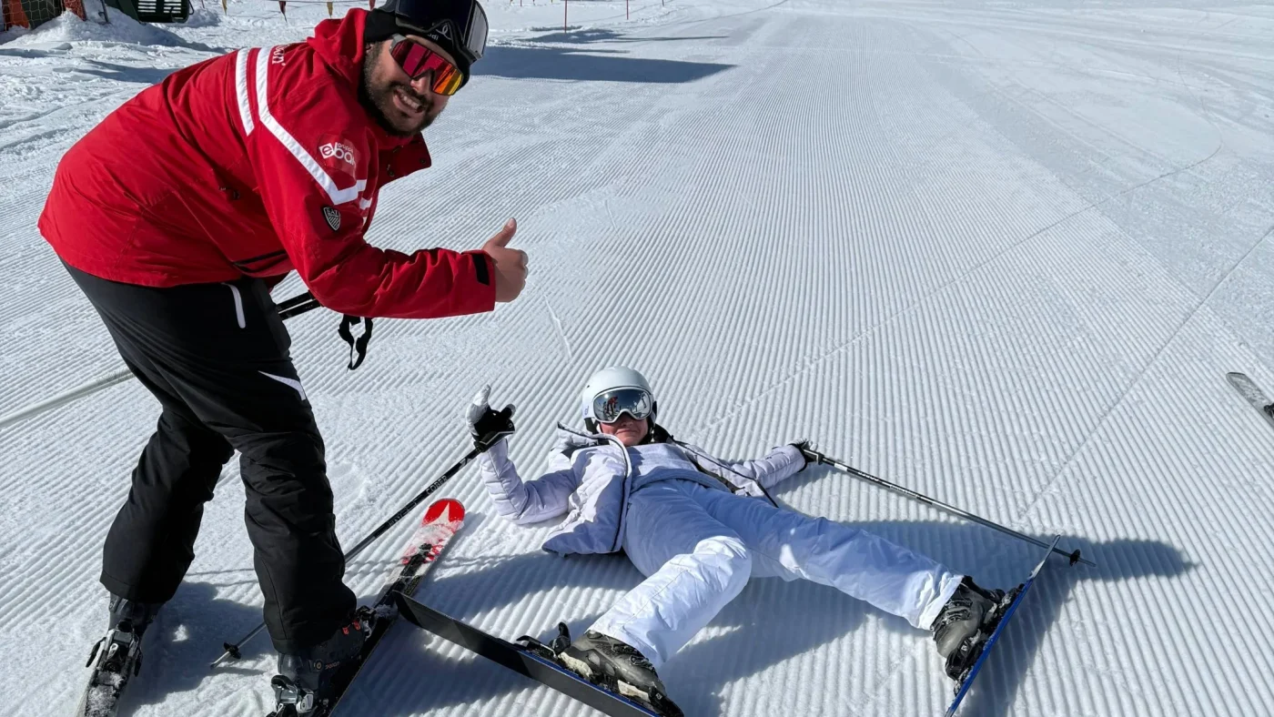 A Barney teacher does a thumbs up next to a student who's fallen while skiing.