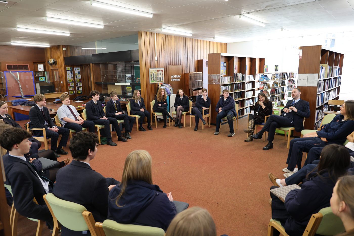 Students and staff sit in a circle in a library space, engaged in a group discussion or workshop.