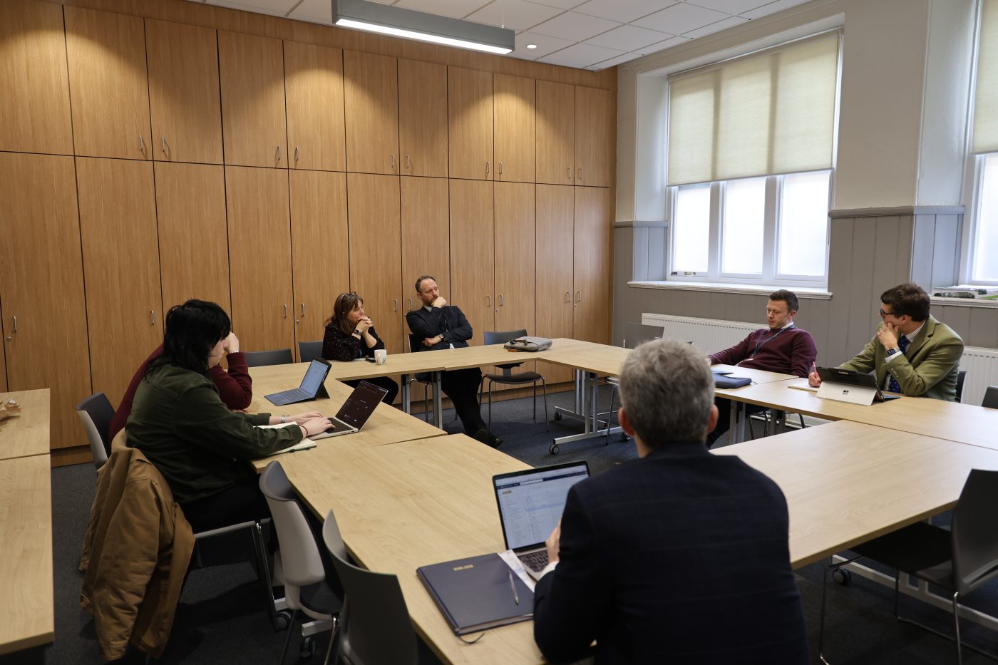 A small group of adults seated around a U-shaped table in a bright meeting room, engaged in discussion, with laptops and notebooks open as one person speaks and others listen attentively.