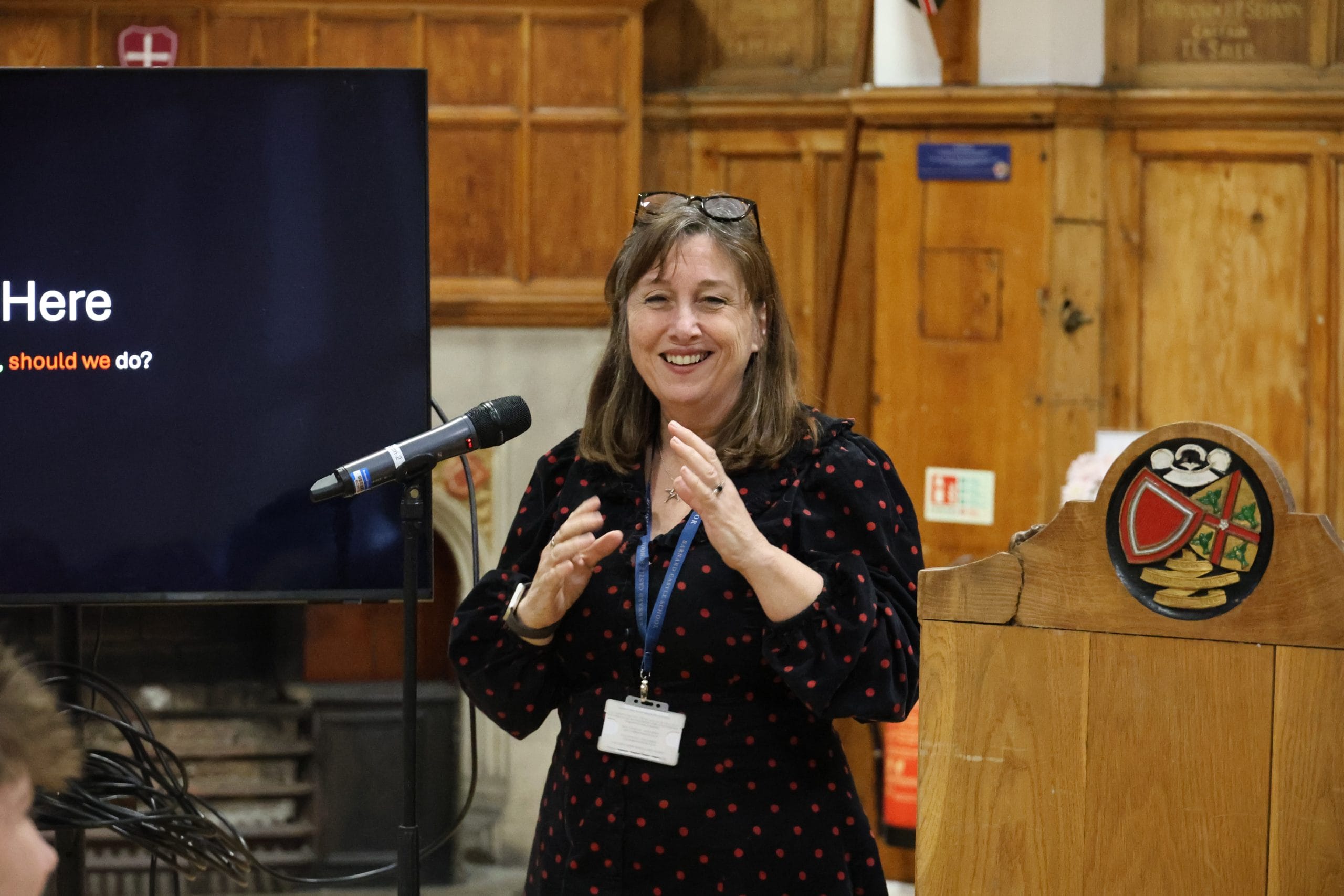 BBC's Laura Ellis stands smiling beside a podium in a wood-panelled hall, speaking into a microphone while delivering a presentation, with a screen displaying part of her talk behind her.