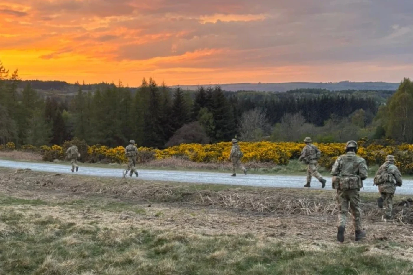 Several students in camouflage gear run across a dirt path during a scenic sunset with a backdrop of trees and yellow flowers.