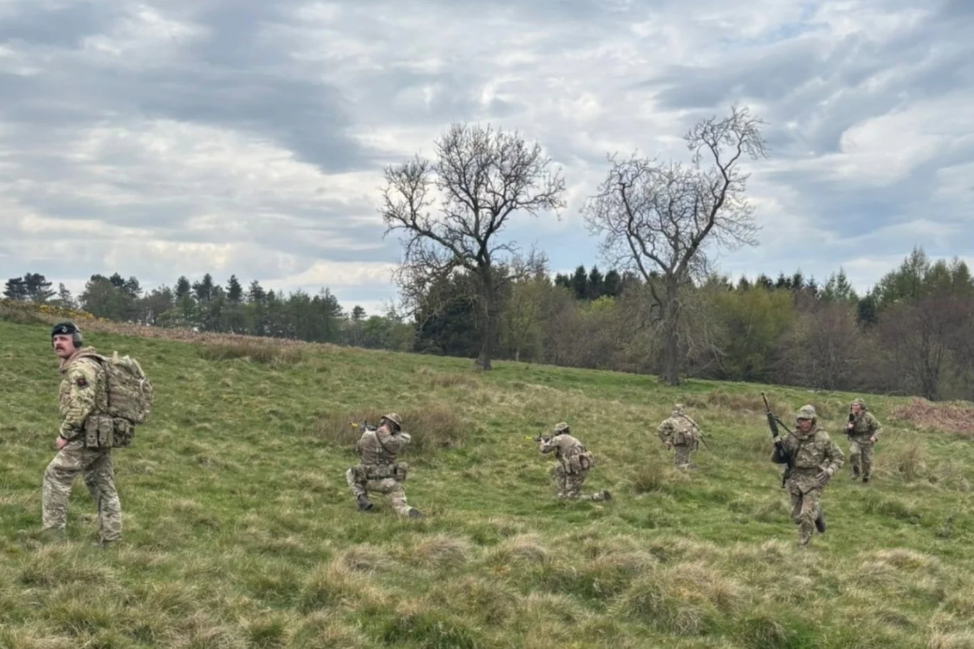 A group of students in camouflage uniforms move across a grassy field, with some kneeling to aim rifles and others advancing under a cloudy sky.