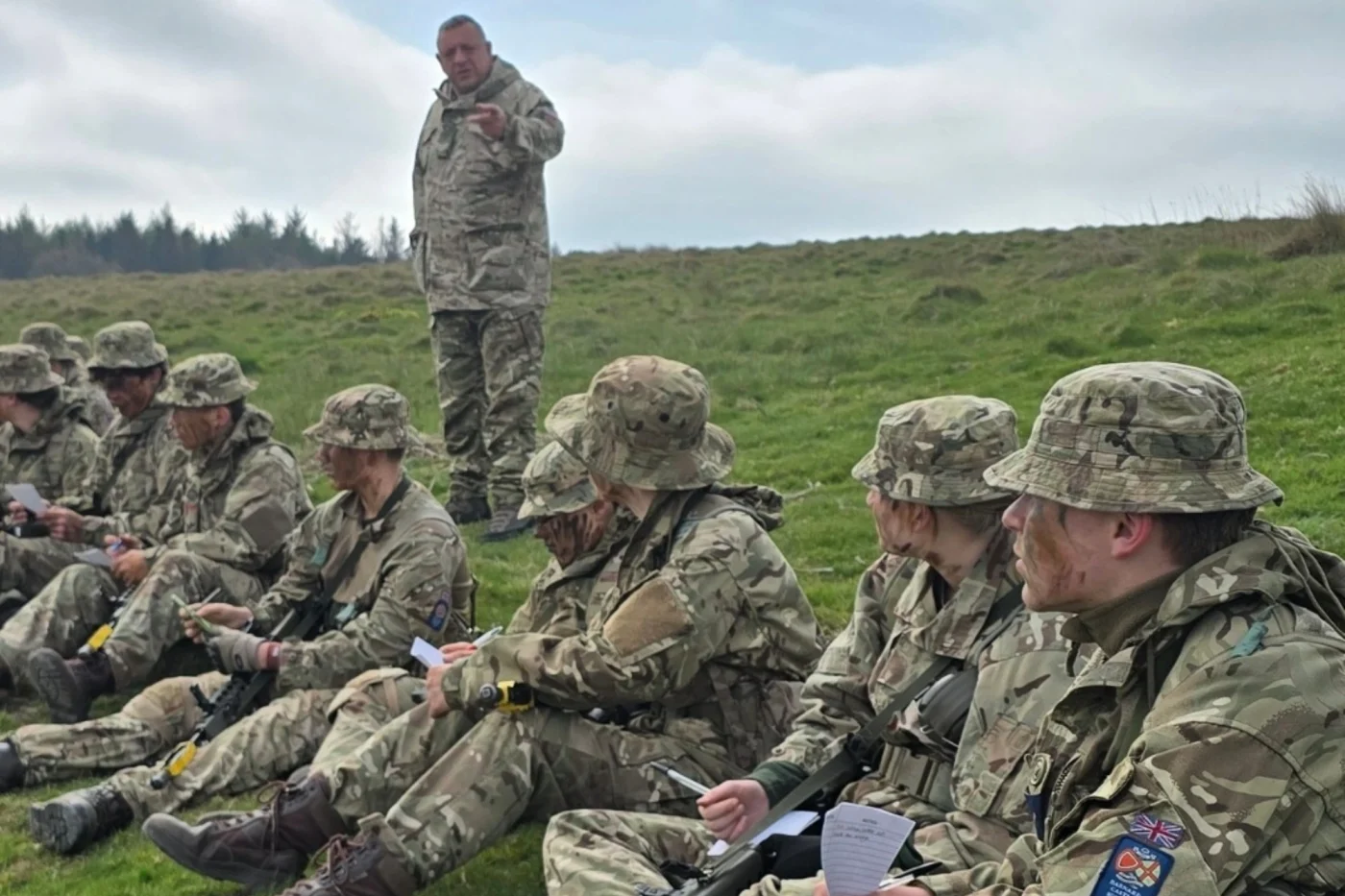 A group of students in camouflage uniforms sit on a grassy hillside taking notes while an instructor stands and speaks to them during a field exercise.