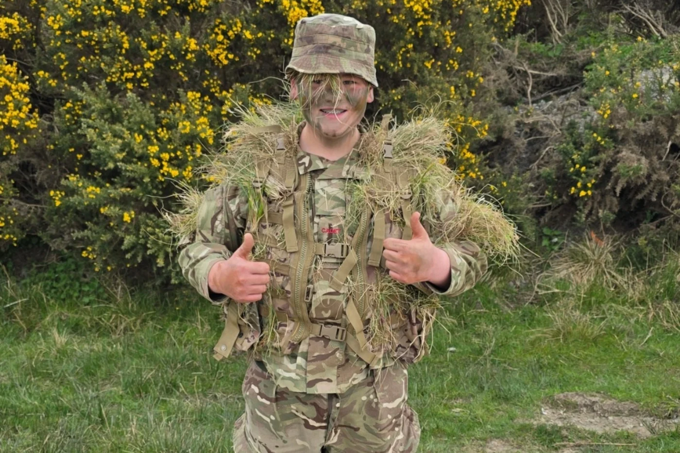 A student in camouflage uniform and face paint gives two thumbs up while wearing grass for extra concealment during a training exercise.