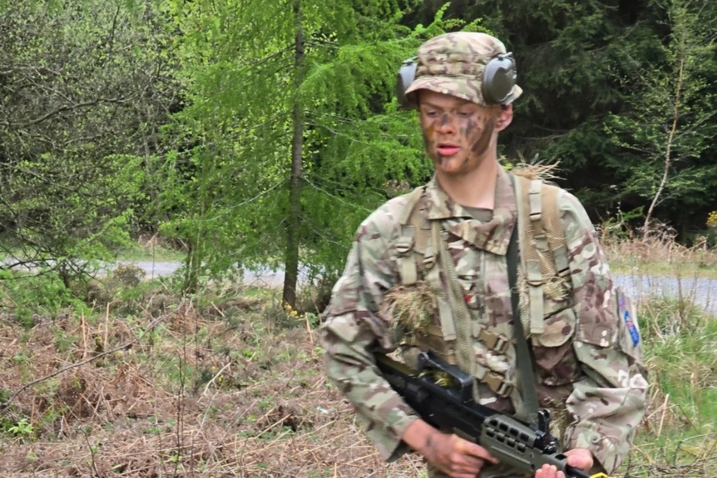 A student in camouflage uniform and face paint walks through a wooded area while carrying a rifle during a training exercise.