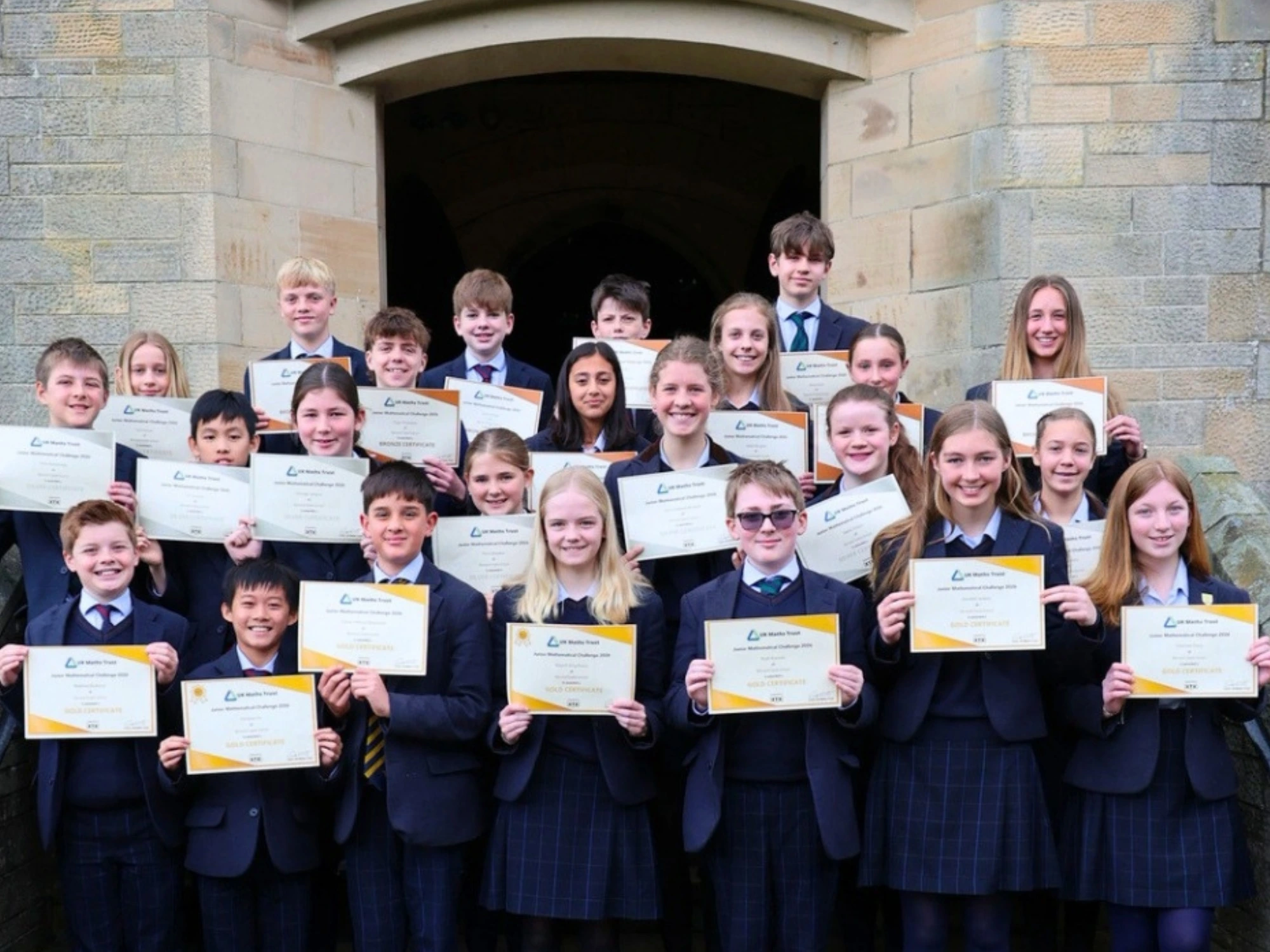 Students stood in stone entranceway smiling and holding up their maths certificates.