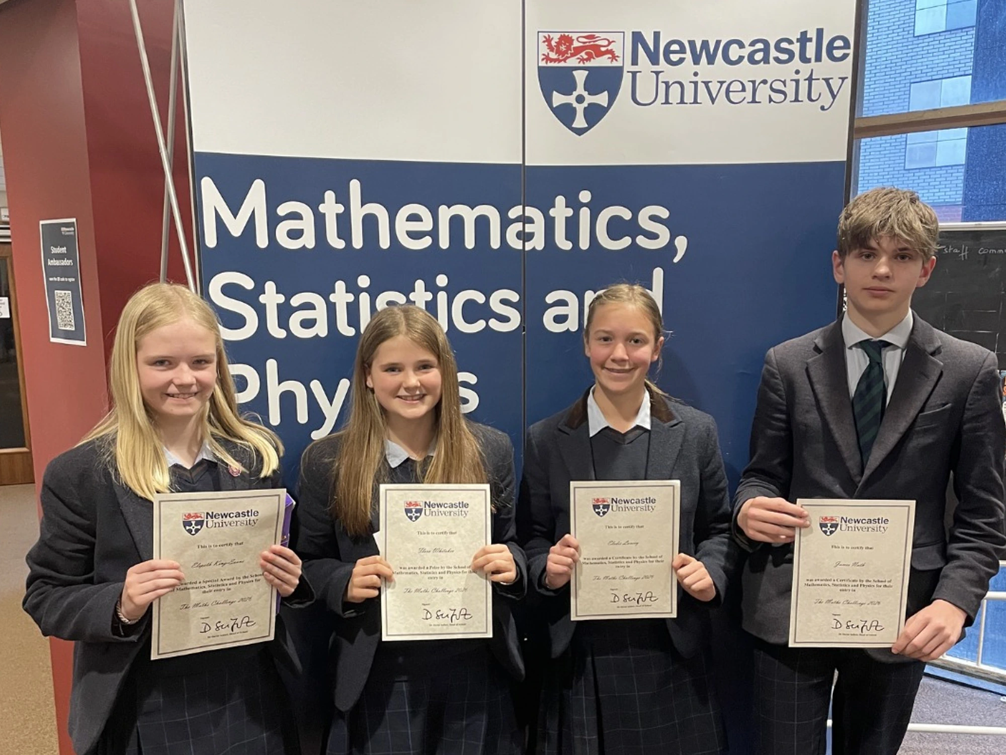 Four pupils stood in front of Newcastle University maths banner holding award certificates for maths competition.