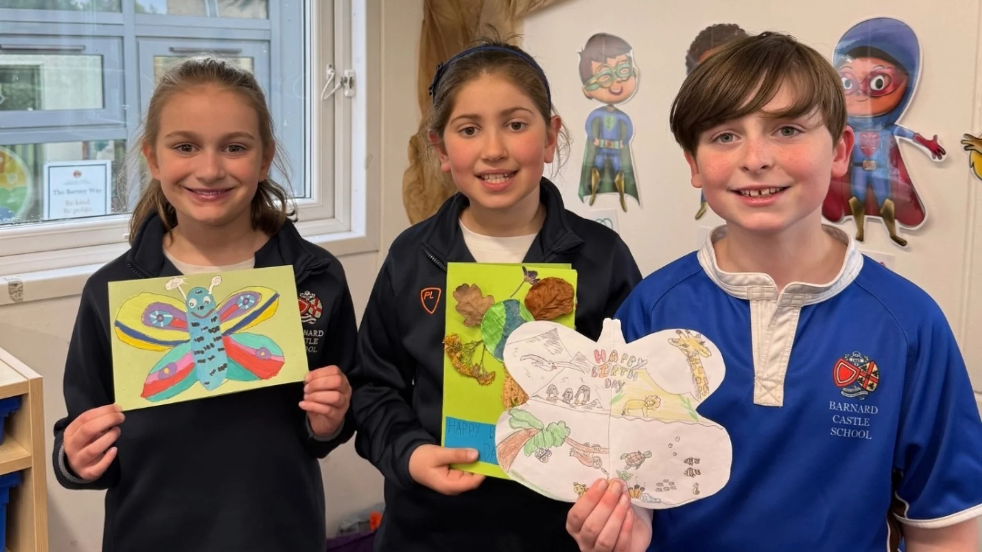 Three Barney pupils showing their hand-made birthday cards for Sir David Attenborough's 100th birthday.
