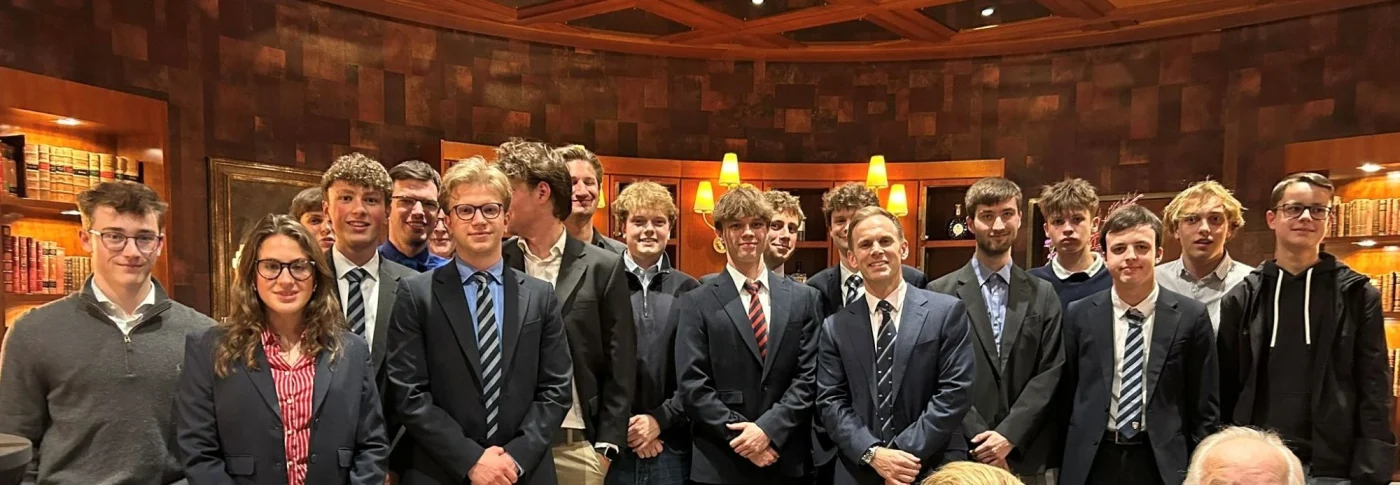 A group of students in formal suits, blazers, and ties pose together inside a room with wood-panelled walls and bookshelves.