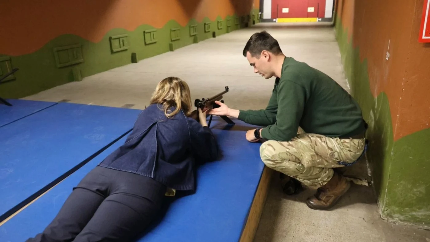 A woman holding a rifle while being instructed by a member of Barney staff.