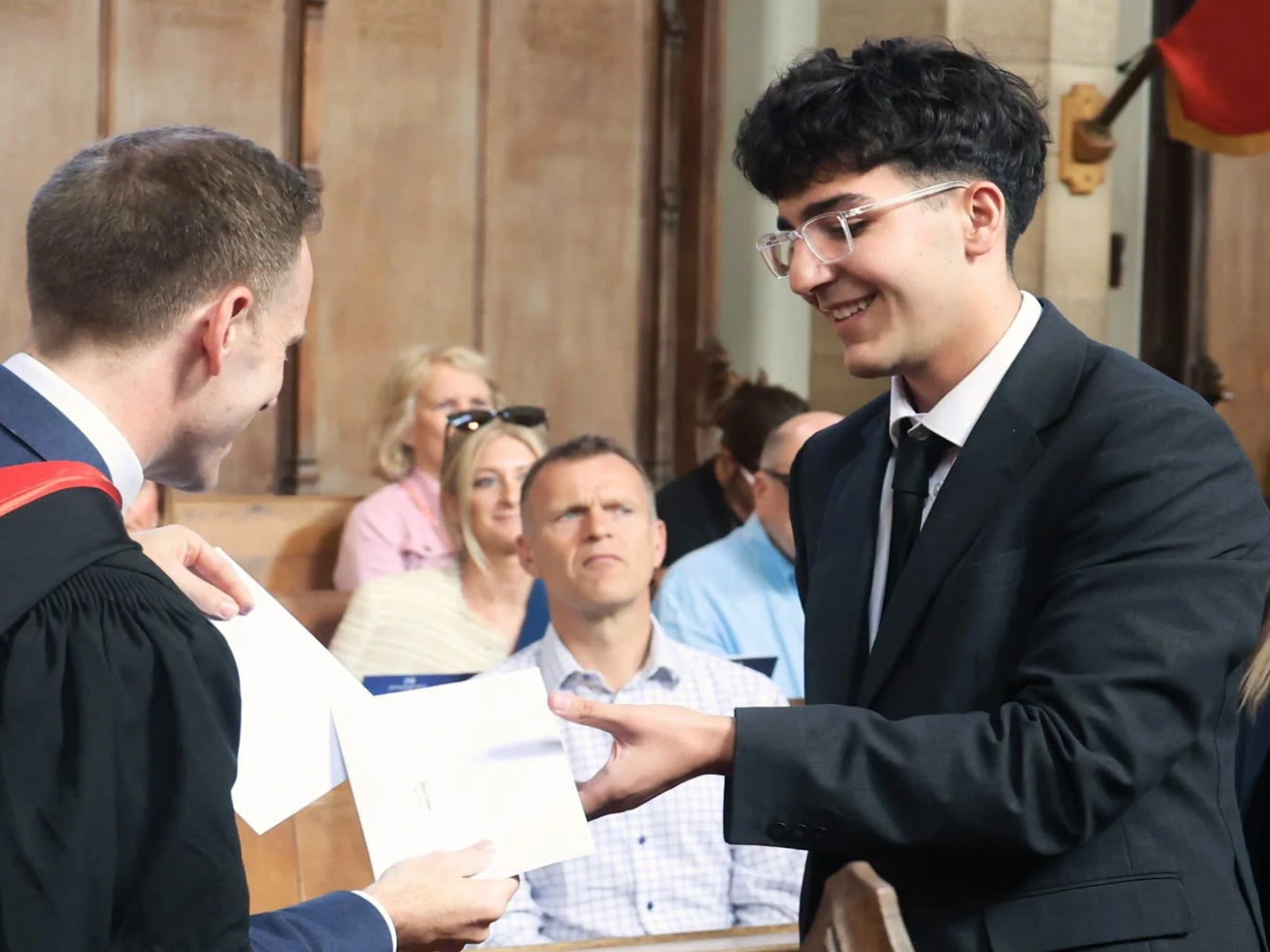 Young man with black hair and glasses receives prize in envelope from headmaster.